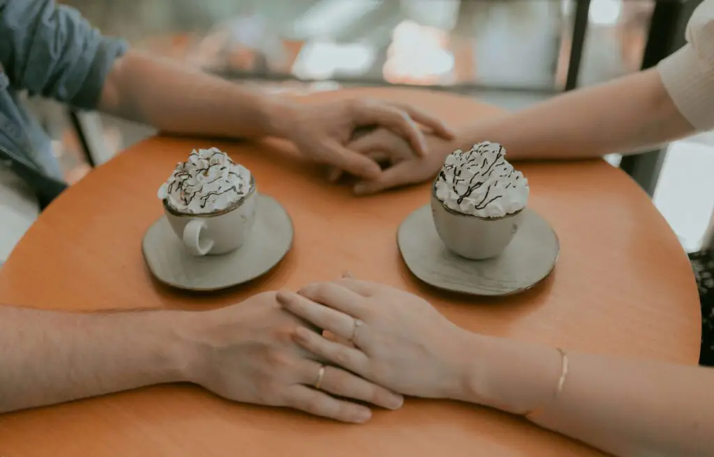 Two cappuccinos on a café table with the hands of a couple in love gently touching across the table.
