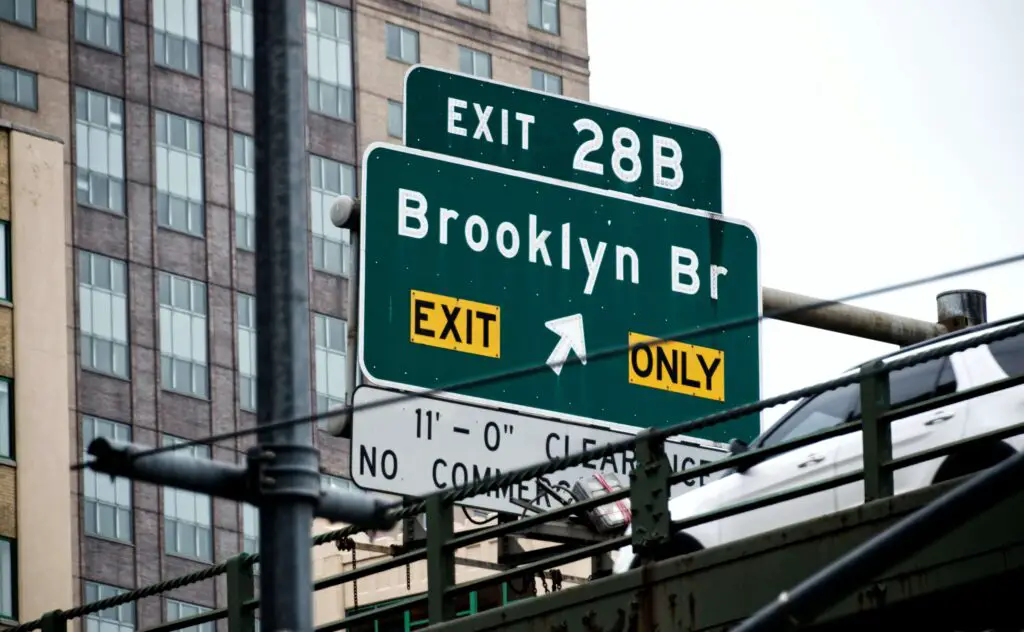 Brooklyn Bound Explorations: Uncovering Urban Gems in New York City Street signs and directional plaques pointing toward Brooklyn, set against the backdrop of a bustling New York City street.