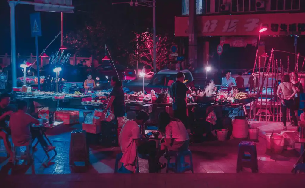 Nightlife and Evening Entertainment in a Different Light: Activities in the Lower East Side People enjoying food and conversation at night in a lively plaza on the Lower East Side, surrounded by warm lights and open-air dining.