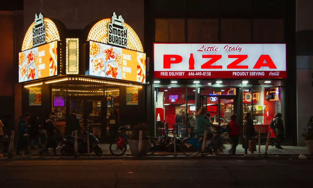 Greenwich Village Food and Nightlife: Best Activities to Do for Tourists Exterior of a cozy pizzeria and a lively burger joint side by side in Greenwich Village, inviting passersby with warm lights and local charm.