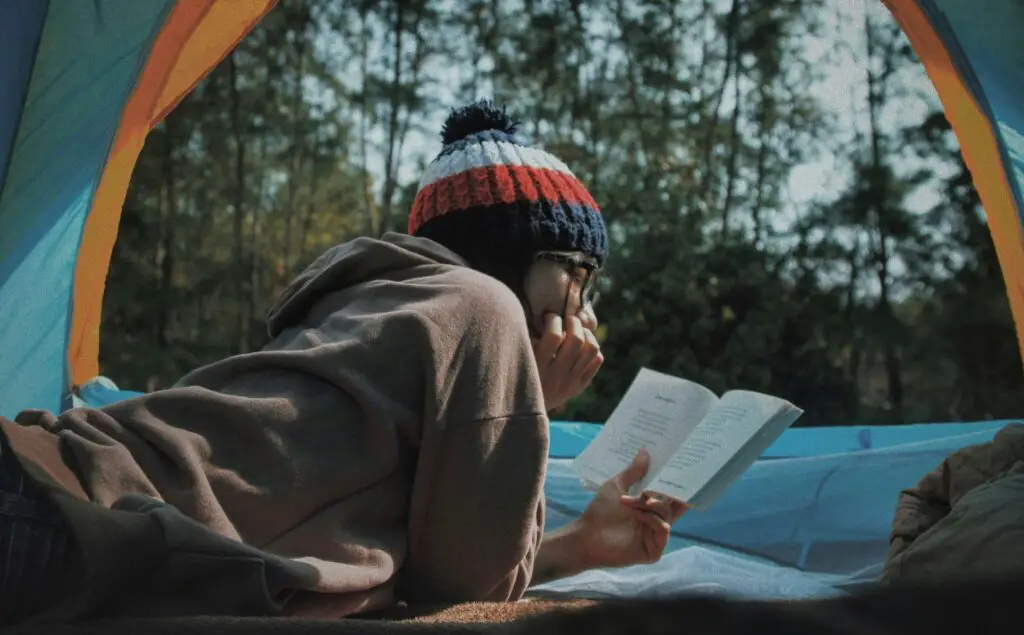 Intellectual Adventures and Curious Pursuits: Indoor Activities in Brooklyn for Adults Young woman reading a book inside her tent, surrounded by trees in a peaceful forest setting.