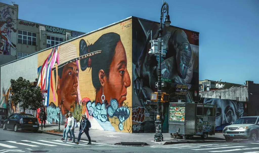 Neighborhood Snapshot and Vibe: Things to Do in Bushwick NYC People crossing the street in Bushwick, NYC, with colorful street art murals decorating the walls behind them.